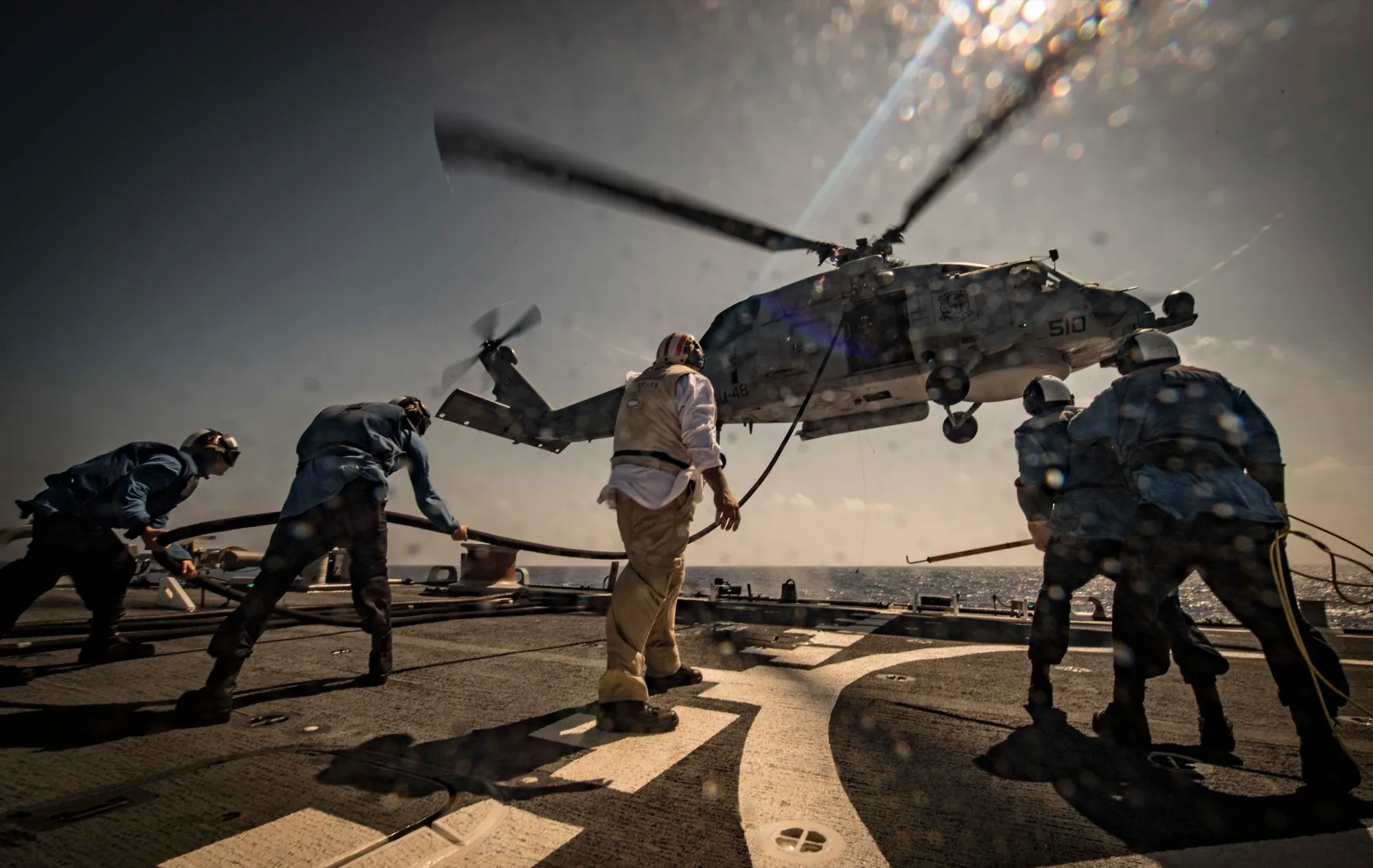 Sailors drag a fuel line beneath an MH-60R Seahawk hovering over a destroyer's flight deck